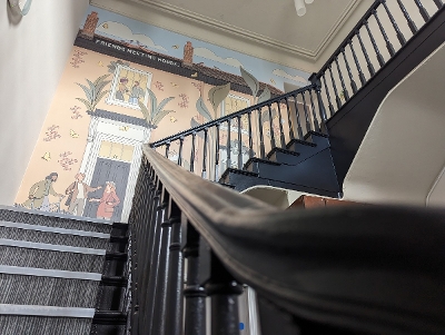 A wooden staircase winding up with a mural of a friends meeting house on the back wall